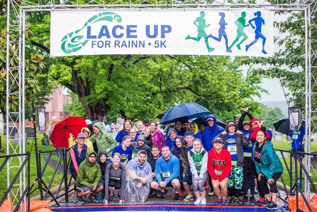 Runners pose in the rain under a banner that reads "Lace Up For RAINN 5k." Part of a RAINN fundraising event.  